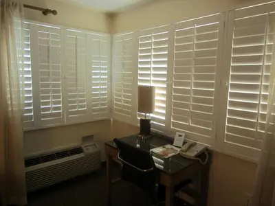 a room with white shuttered windows and a table with a lamp at PB Surf Beachside Inn in San Diego