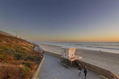 two people walking down a sidewalk next to the beach at Luxury Ocean Views & Steps To The Sand - Tower 36 Upper Condo Unit in Carlsbad